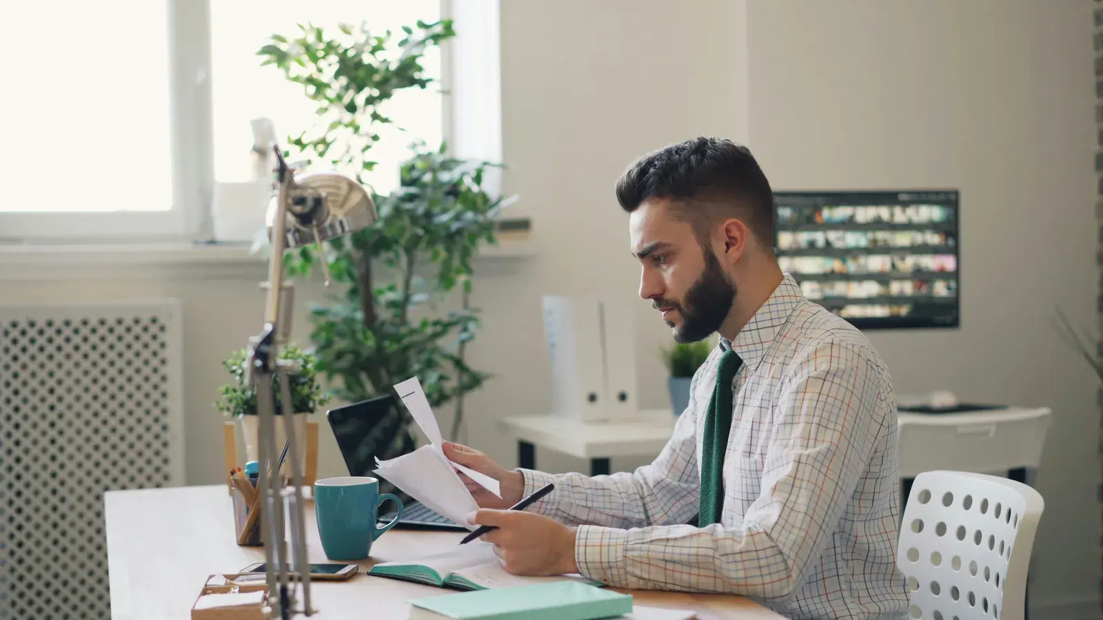 A man at a desk in a bright office reading printed papers with a pen in hand